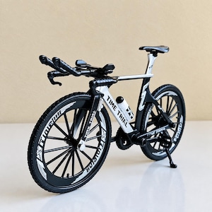 May include: A miniature black and white time trial bicycle model featuring aerodynamic handlebars, a sleek frame with the text TIME TRIAL printed on the side, thin spoked wheels with white lettering, a small water bottle, and a kickstand.