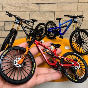 Könnte beinhalten: Drei Miniatur-Mountainbike-Modelle in Rot, Blau und Gold, jeweils mit schwarzen Rädern, einem gefederten Rahmen und einem kleinen Nummernschild mit der Aufschrift 31 am Lenker. Die Rahmen tragen die Aufschrift DOWN HILL und FORK.