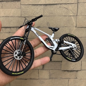 Può includere: Un modellino di mountain bike bianco con ruote nere e scritte gialle, sella nera, sospensione posteriore e manubrio nero. Il telaio riporta le scritte 08 e HARD, mentre la forcella anteriore è contrassegnata con la scritta FORK.