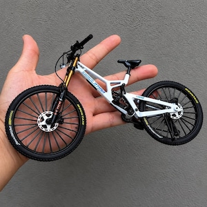 Peut inclure: Un modèle réduit de VTT blanc avec des pneus noirs à lettrage jaune, des fourches de suspension avant dorées, une selle noire et un cadre portant l'inscription SIMULATION. Le vélo présente des rayons détaillés et des freins à disque.