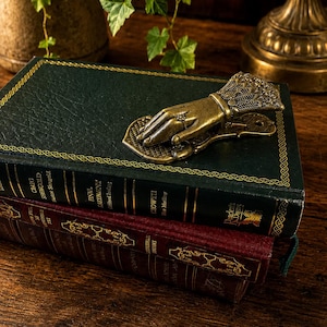 Puede incluir: Un clip de metal color latón con forma de mano sobre un libro de tapa dura verde oscuro. El clip presenta detalles de filigrana en el puño y un pequeño anillo en el dedo. El libro está apilado sobre un segundo libro de color granate.