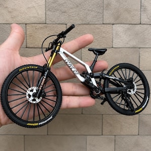 Peut inclure: Un modèle réduit de vélo tout-terrain avec un cadre argenté portant l'inscription COME ON, des roues noires à pneus crantés, des fourches de suspension avant dorées et une selle noire. Le vélo présente des rayons, des freins à disque et une chaîne.