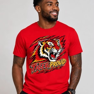 May include: A bright red short-sleeved crew neck t-shirt featuring a graphic of a roaring tiger head surrounded by claw marks. Below the tiger, the text TIGER PROUD is printed in a bold, stylized yellow and black font with a distressed effect.