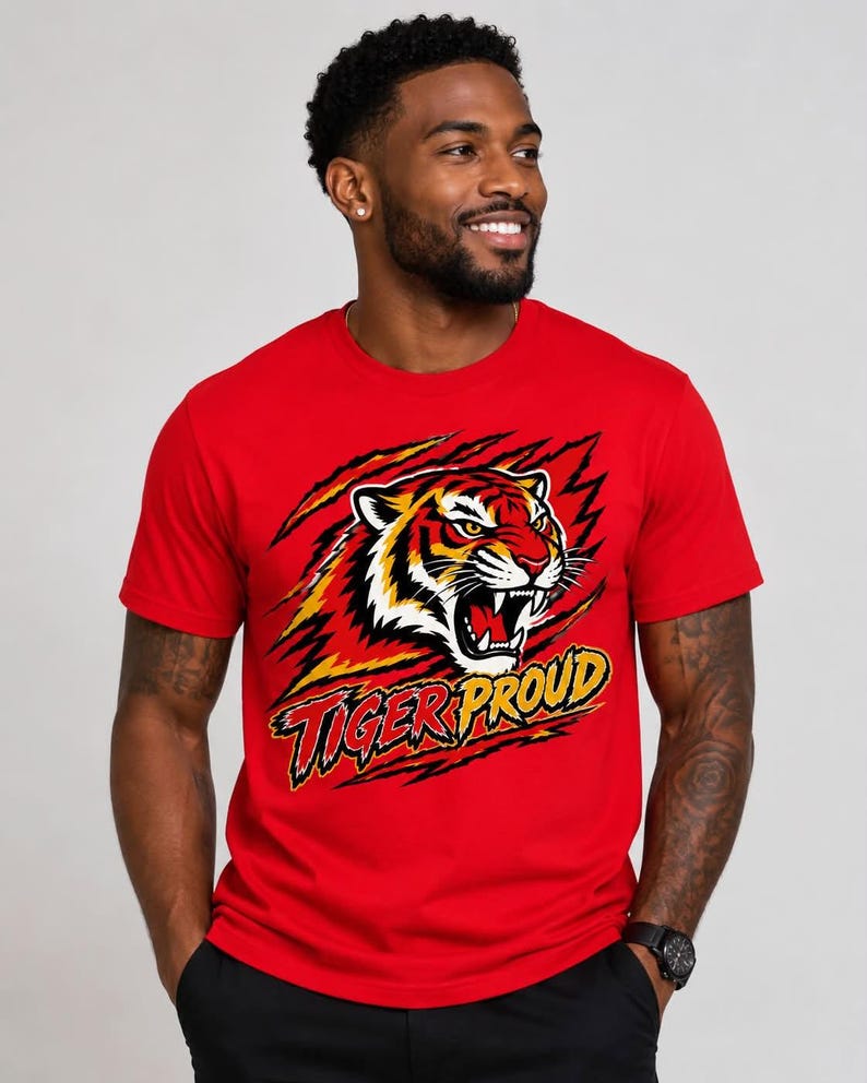 May include: A bright red short-sleeved crew neck t-shirt featuring a graphic of a roaring tiger head surrounded by claw marks. Below the tiger, the text TIGER PROUD is printed in a bold, stylized yellow and black font with a distressed effect.