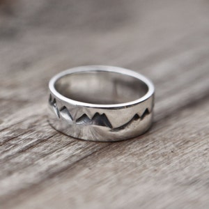 Puede incluir: Un anillo de plata con un dise&ntilde;o de cordillera. El anillo est&aacute; sobre una superficie de madera.