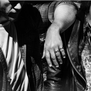 Puede incluir: Dos personas de pie cerca una de la otra, una con una camisa a cuadros y la otra con una chaqueta de cuero. La persona con la camisa a cuadros tiene el brazo alrededor del hombro de la otra persona. La persona con la chaqueta de cuero lleva un anillo en el dedo.