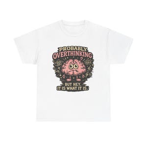 Pode incluir: Uma t-shirt branca com um gráfico de um cérebro cor-de-rosa com aspeto preocupado, com braços e pernas. O texto acima diz PROBABLY OVERTHINKING em cor-de-rosa, e o texto abaixo diz BUT HEY, IT IS WHAT IT IS a preto, rodeado de rabiscos.