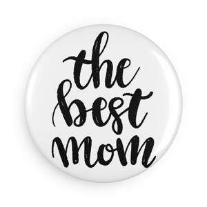 May include: A circular white pin-back button featuring the black handwritten-style text "the best mom" centered on the surface. The design uses a bold, brush-stroke font against a plain, solid white background, creating a high-contrast appearance.