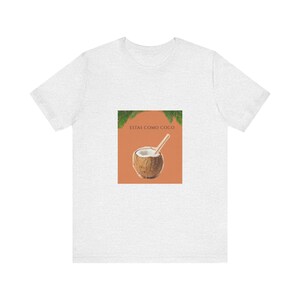 May include: A light gray t-shirt featuring a central rectangular graphic on the chest. The graphic displays a brown coconut with a straw, green palm leaves at the top corners, and the text "ESTAS COMO COCO" printed in dark brown letters.