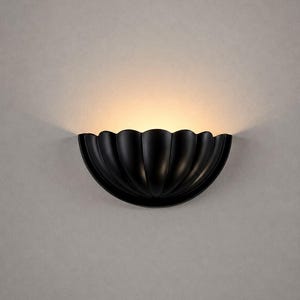 May include: A matte black wall sconce featuring a semi-circular, scalloped shell design. The fixture emits a warm, soft glow from its top edge, casting light upward against the wall surface to create a focused and ambient illumination effect.