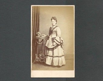 Könnte beinhalten: Ein Vintage-Foto in Sepia-Ton einer Frau in einem langen Kleid und Jacke, die neben einem kleinen Tisch steht. Das Kleid hat einen geschichteten Rock und die Jacke hat dekorative Verzierungen. Der Tisch hat verzierte Details und ein kleines Blumenarrangement.