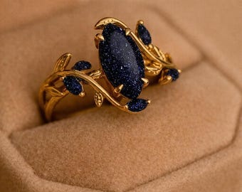 Könnte beinhalten: Ein goldener Ring mit einem zentralen, marquise-geschliffenen dunkelblauen Sandstein mit schimmernden Sprenkeln. Die Ringschiene ist mit zarten goldenen Metallblättern und kleinen, ovalen blauen Sandsteinakzenten in der Struktur gestaltet.