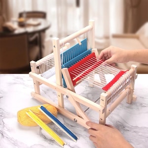 May include: A wooden tabletop loom featuring a blue heddle and a red heddle with white warp threads strung through them. Beside the loom sits a ball of yellow yarn, a yellow shuttle with yellow thread, and a blue shuttle with blue thread.
