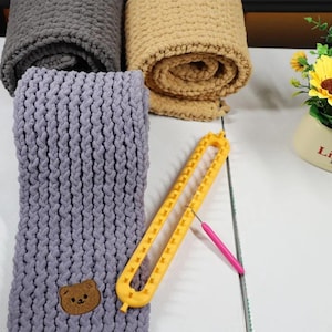 Könnte beinhalten: Ein lavendelfarbener, grob gestrickter Schal mit einem kleinen braunen Bärenaufnäher, neben einem gelben Strickrahmen aus Kunststoff und einem rosa Häkelhaken. Im Hintergrund sind zwei weitere gerollte, grob gestrickte Decken in Grau und Beige.