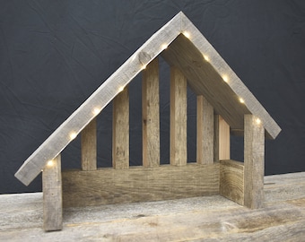 Peut inclure: Une crèche de Noël rustique en bois avec un toit triangulaire et une guirlande lumineuse qui souligne la structure. La scène est faite de bois vieilli et a un aspect naturel et rustique.