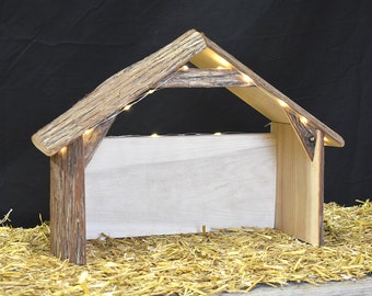 Peut inclure: Une crèche de Noël en bois avec une finition rustique brun et blanc. La scène présente un petit abri ouvert sur les côtés avec un intérieur blanc et un toit en bois avec une texture d'écorce. L'abri est éclairé par une guirlande de lumières blanches chaudes.