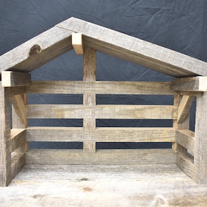 Könnte beinhalten: Ein rustikaler Krippenstall aus Holz mit Satteldach. Die Struktur besteht aus verwittertem, grauem Holz mit Latten an der Vorder- und Seitenwand. Das Design erinnert an Einfachheit und traditionelle Handwerkskunst.