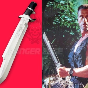 May include: A large survival knife with a silver-toned steel blade and a black handle featuring a prominent crossguard. The blade has a distinct clip point design and a serrated spine. The knife is shown against a bright red background.