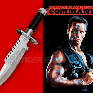 May include: A silver-colored survival knife with a black handle and serrated spine, positioned next to a promotional image for the film Commando featuring Arnold Schwarzenegger. The blade includes engraved text and a metal crossguard.