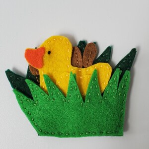 May include: A felt applique featuring a yellow duck with an orange beak and brown wings, nestled within a patch of bright green grass. The design is constructed from layered felt pieces with visible stitching details along the edges of the grass.