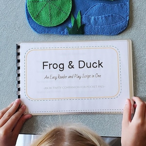 Könnte beinhalten: Eine spiralgebundene Broschüre mit dem Titel Frog & Duck: An Easy Reader and Play Script in One und dem Untertitel An Activity Companion for Pocket Pals. Über der Broschüre liegt eine blaue Filzmatte mit einem grünen Frosch auf einem Seerosenblatt.
