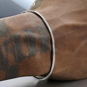 Peut inclure: Un bracelet chaîne à motif chevrons de couleur argentée, doté d'un design à maillons plats, flexibles et étroitement tissés qui repose sur la peau. La surface métallique reflète la lumière, révélant une texture lisse et uniforme.