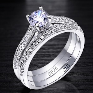 May include: A silver-toned ring set featuring a central round-cut clear stone held in a four-prong setting. The band and the accompanying ring are encrusted with small clear stones along the top surface. The inner band is engraved with S925.