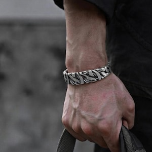 Puede incluir: Un brazalete de metal en tono plateado con un diseño texturizado, orgánico y superpuesto, con surcos profundos y un acabado pulido. El brazalete tiene una estructura de banda ancha y rígida que rodea la muñeca, mostrando un patrón complejo e irregular.