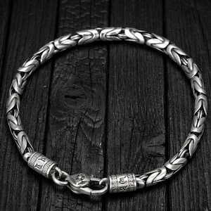 Pode incluir: Uma pulseira de corrente de metal prateado com um padrão de elos bizantinos entrelaçados. A pulseira inclui duas tampas cilíndricas gravadas com símbolos e um fecho lagosta que conecta as duas extremidades da corrente circular.