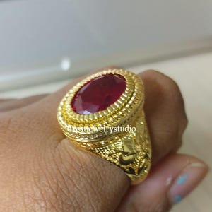 May include: A gold ring featuring a large, oval-cut, deep red gemstone set in a textured, rope-style bezel. The band is wide and decorated with intricate, embossed floral patterns. The text "onsthewelrystudio" is visible on the ring surface.