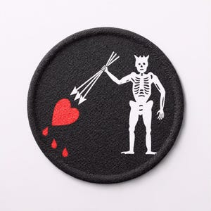 以下が含まれることがあります： 黒い円形の刺繍ワッペン。白い骸骨が3本の白い矢を持ち、赤いハートに向けている様子が描かれている。ハートはワッペンの左側に配置され、下端からは3つの赤い雫が滴り落ちているデザインとなっている。