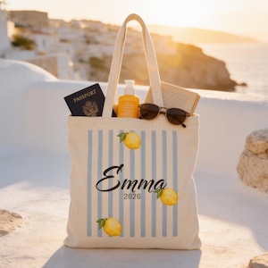 May include: A cream-coloured canvas tote bag featuring vertical light blue stripes, yellow lemon illustrations, and the printed text "Emma 2026". The bag contains a dark blue passport, a yellow bottle, a pair of sunglasses, and an open book.