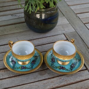 Może przedstawiać: Dwie turkusowe filiżanki do herbaty z porcelany ze złotymi obw&oacute;dkami i kwiatowymi wzorami na pasujących spodkach, ustawione przed ciemną ceramiczną doniczką z kwitnącą rośliną o licznych jasnor&oacute;żowych kwiatach z ż&oacute;łtymi środkami.