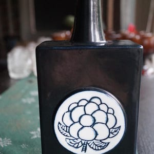 Op de afbeelding: Een rechthoekige zwarte keramische fles met een smalle hals. Centraal op de voorzijde bevindt zich een wit cirkelvormig medaillon met een blauwe lijntekening van een wolkbes. Een klein label onderaan bevat de tekst ARABIA handicraft.