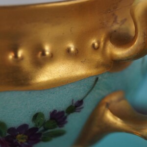 Może przedstawiać: Porcelanowy kubek z szerokim złotym brzegiem ozdobionym wypukłymi kropkami. Korpus kubka ma jasny turkusowy kolor i jest ozdobiony ręcznie malowanymi fioletowymi kwiatami oraz zielonymi liśćmi. Z boku znajduje się złoty uchwyt.