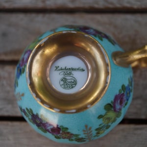 Może przedstawiać: Jasnoniebieska filiżanka porcelanowa z kwiatowym wzorem w kolorze fioletowym, r&oacute;żowym i zielonym. Podstawa jest wykończona złotem, a na spodzie znajduje się zielona pieczątka z napisem Hutschenreuther Selb oraz symbolem lwa.