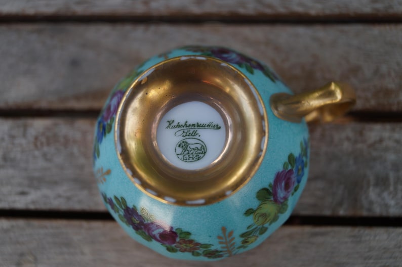 Może przedstawiać: Jasnoniebieska filiżanka porcelanowa z kwiatowym wzorem w kolorze fioletowym, r&oacute;żowym i zielonym. Podstawa jest wykończona złotem, a na spodzie znajduje się zielona pieczątka z napisem Hutschenreuther Selb oraz symbolem lwa.