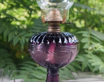 Może przedstawiać: Fioletowa szklana lampa naftowa z teksturowanym, pękatym zbiornikiem i pasującą podstawą z misternymi geometrycznymi wzorami. Lampa posiada mosiężny palnik oraz przezroczysty, zwężający się ku górze szklany komin.