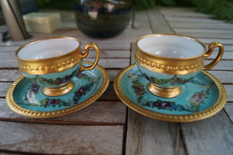 Może przedstawiać: Dwie pasujące filiżanki do herbaty ze spodkami z porcelany, o jasnoniebieskim tle z ręcznie malowanymi girlandami kwiatowymi w odcieniach fioletu, r&oacute;żu i zieleni. Zestaw posiada złocone krawędzie, uszka i podstawy o perłowej fakturze.