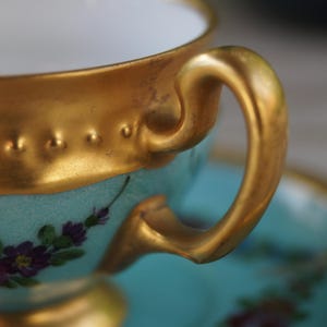 Może przedstawiać: Jasnoniebieska filiżanka do herbaty z porcelany ze złoconym brzegiem o wypukłej, koralikowej fakturze. Filiżanka posiada zakrzywione złote ucho i jest ozdobiona małymi, malowanymi fioletowymi i r&oacute;żowymi kwiatami. Widoczny jest pasujący spodek.