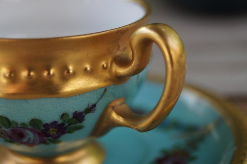 Może przedstawiać: Jasnoniebieska filiżanka do herbaty z porcelany ze złoconym brzegiem o wypukłej, koralikowej fakturze. Filiżanka posiada zakrzywione złote ucho i jest ozdobiona małymi, malowanymi fioletowymi i r&oacute;żowymi kwiatami. Widoczny jest pasujący spodek.