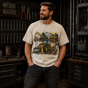 May include: A beige short-sleeve t-shirt featuring a graphic print of yellow construction machinery, including an excavator and a bulldozer, set against a backdrop of mountains, trees, and a yellow sun. The shirt has a crew neckline and a relaxed fit.