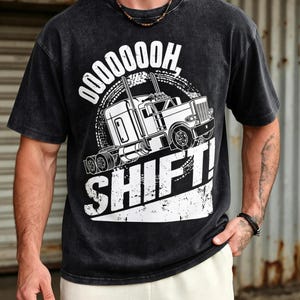 May include: A black acid-wash t-shirt featuring a white graphic of a semi-truck with the text OOOOOOOH, SHIFT! printed above and below the vehicle. The design includes a circular tire tread pattern behind the truck and a distressed white background.
