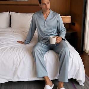 May include: A light blue satin pajama set consisting of a long-sleeved button-down shirt with a notched collar and matching long trousers, paired with white quilted slip-on slippers and a plain white ceramic mug held in one hand.