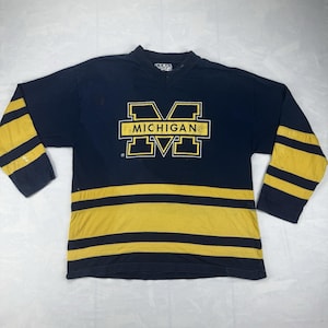 Peut inclure: Un maillot à manches longues bleu marine arborant un grand logo M jaune en bloc avec le mot MICHIGAN imprimé au centre. Les manches et le bas du torse présentent des rayures horizontales jaunes. L'encolure est en V avec une petite étiquette blanche.