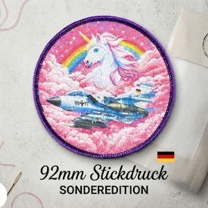 Könnte beinhalten: Ein kreisförmiger Stickaufnäher mit einem weißen Einhorn mit goldenem Horn über einem blau-weißen Kampfjet. Der Hintergrund besteht aus rosa Wolken und einem Regenbogen. Der Aufnäher hat einen violetten Rand und misst 92 mm.