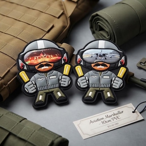 Könnte beinhalten: Zwei 10 cm große PVC-Aufnäher, die einen Flugzeug-Einwinker als Cartoon-Figur zeigen. Die Figur trägt eine graue Jacke, eine olivfarbene Hose und einen Helm mit einem Visier, das einen Jet reflektiert. Jede Figur hält zwei gelbe Stäbe. Ein Etikett trägt die Aufschrift Aviation Marshaller - 10cm PVC.