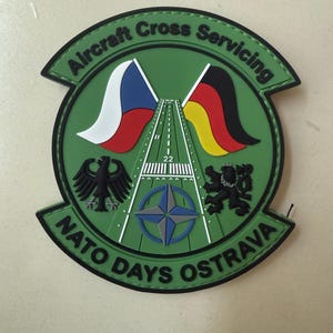Könnte beinhalten: Ein kreisförmiger grüner Aufnäher mit dem Text Aircraft Cross Servicing und NATO DAYS OSTRAVA. Das Design zeigt eine zentrale Landebahn mit der Markierung 22, die Flaggen Tschechiens und Deutschlands, einen schwarzen Adler, einen Löwen und einen NATO-Stern.