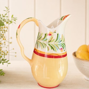 May include: A ceramic pitcher featuring a pale yellow base, a red horizontal band, and a white upper section decorated with hand-painted green leafy sprigs and small red accents. The pitcher has a curved handle and a flared pouring spout.