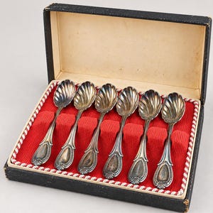 May include: A set of six silver-toned spoons with shell-shaped bowls and decorative handles, arranged in a black rectangular presentation box with a red fabric interior lining and a white braided trim along the inner edge of the box base.